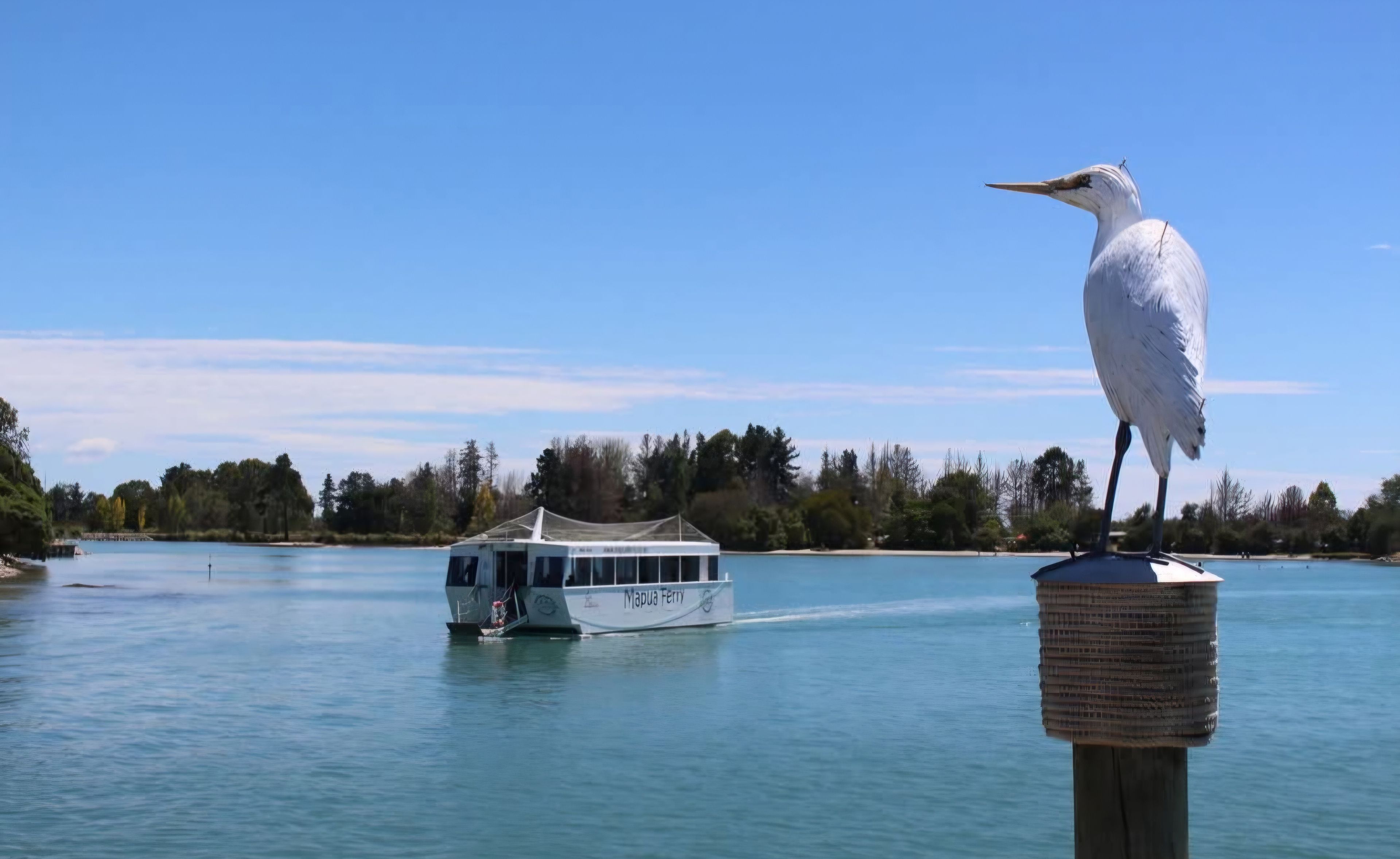 Un ave posada en un poste junto a un lago tranquilo, con un ferry al fondo.