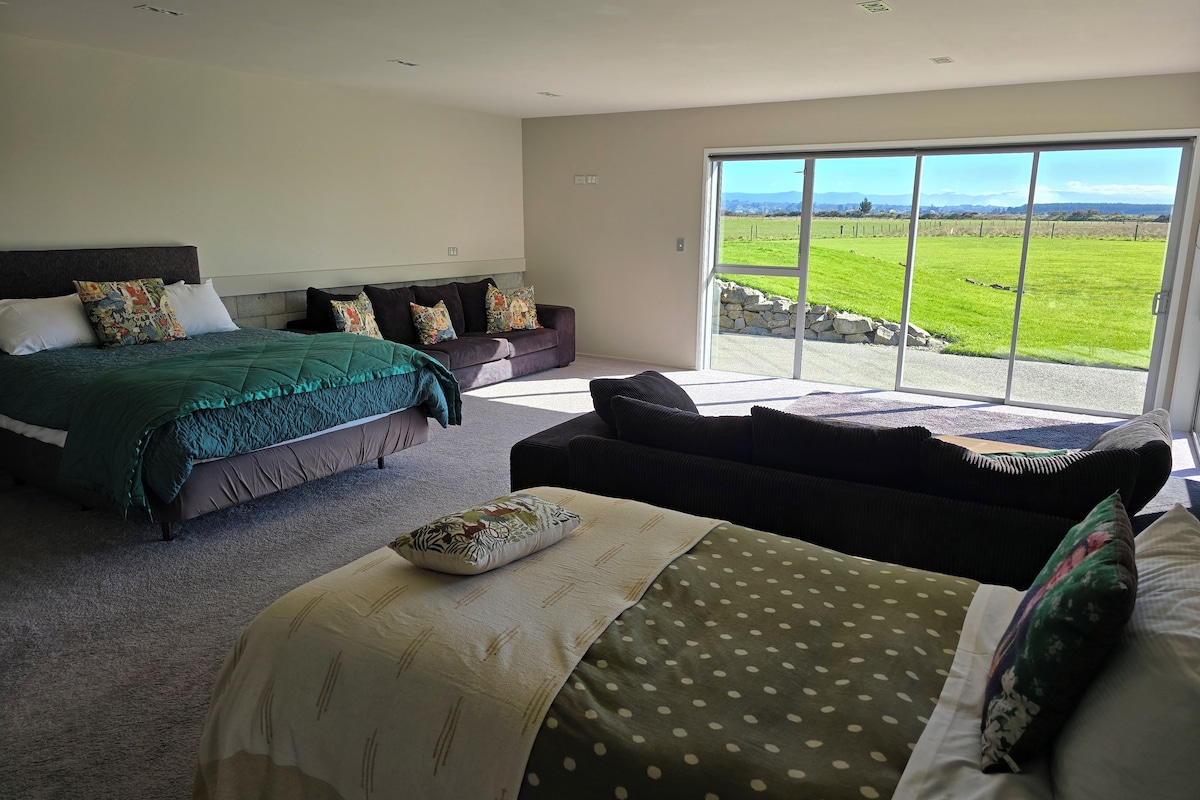 A bedroom with a large bed, a sofa, and a view of a green field through a sliding glass door.