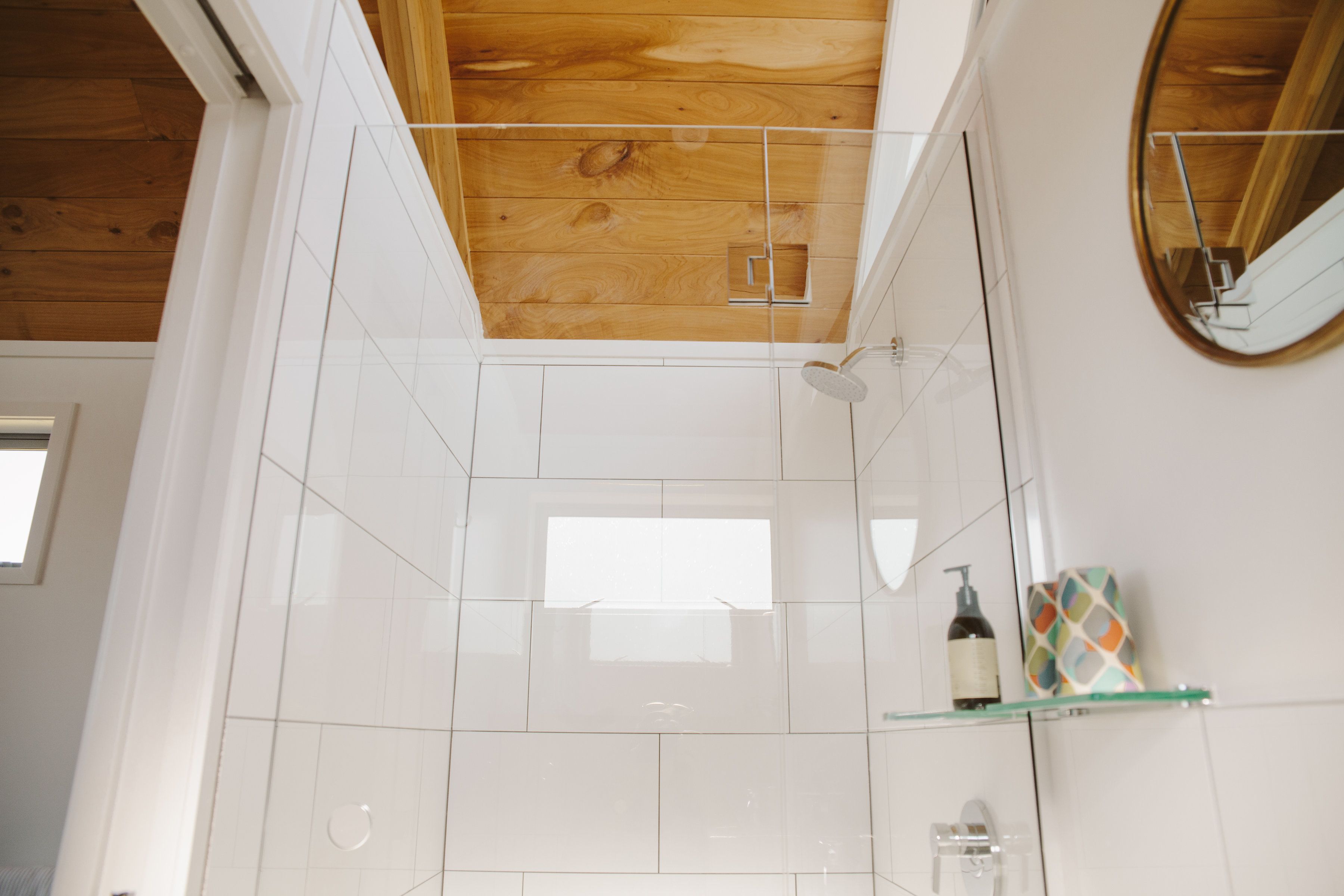 A modern bathroom with a wooden ceiling, white tiles, a glass shower enclosure, and a mirror.