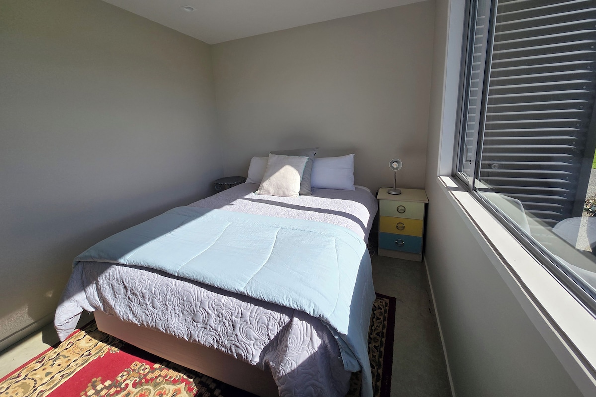 A bedroom with a bed, a nightstand, a bedside table, and a window with blinds.