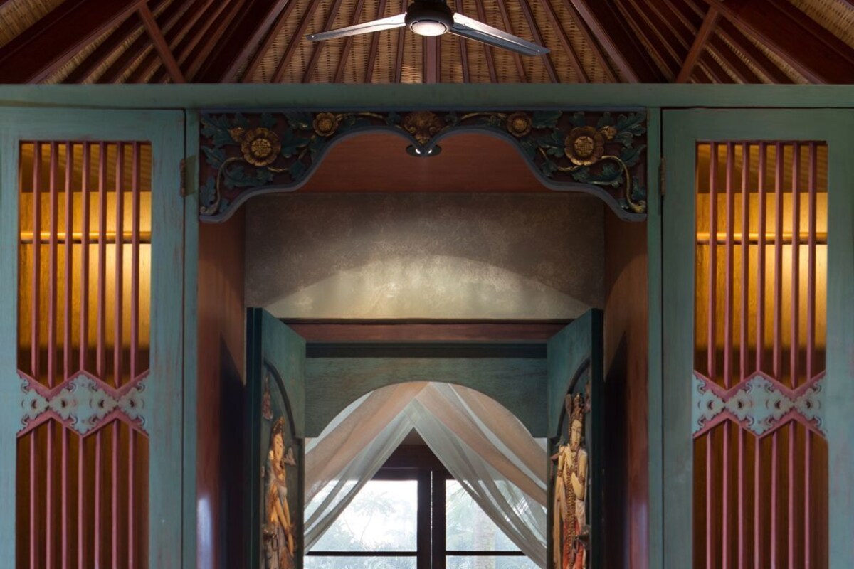 A traditional room with a sloped ceiling, a bed with a headboard, a small table, and a window with a view of greenery.