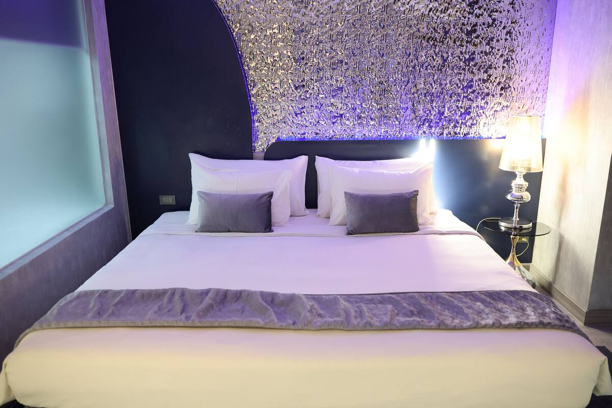 Una cama bien hecha con ropa de cama blanca y gris, una pared decorativa con un patrón texturizado y una mesita de noche con una lámpara al lado.