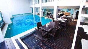 An indoor swimming pool with a tiled floor, surrounded by dark wooden deck and wicker furniture. - Glacier Hotel Khon Kaen (Khon Kaen)