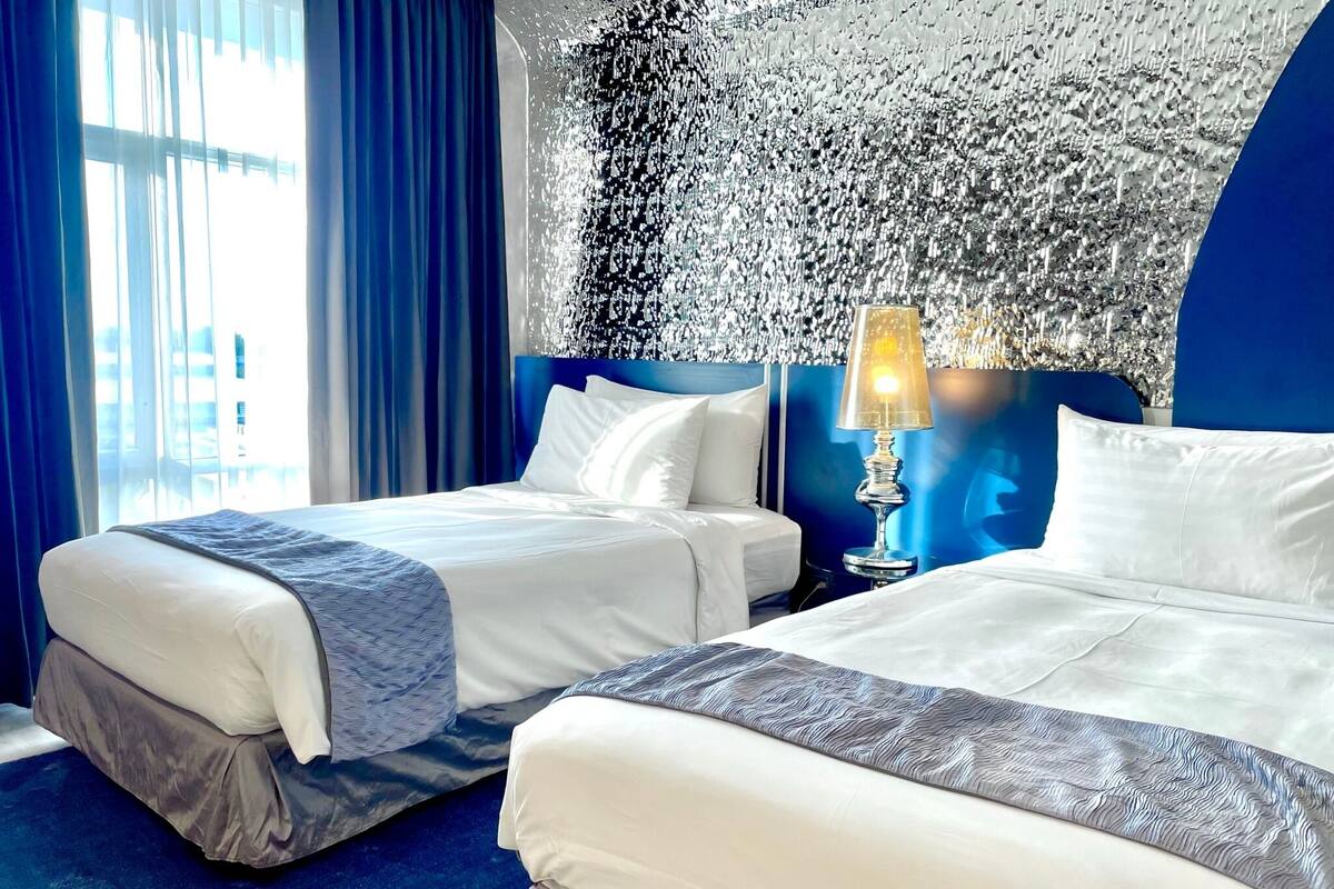 Habitación de hotel con dos camas, un ventanal grande con cortinas, un muro decorativo y una mesita de noche con lámpara.