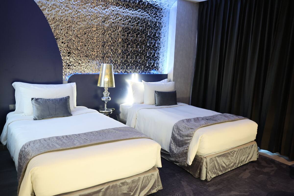 Habitación de hotel con dos camas, un mural grande en la pared, una mesita de noche con lámpara y una ventana con cortinas.