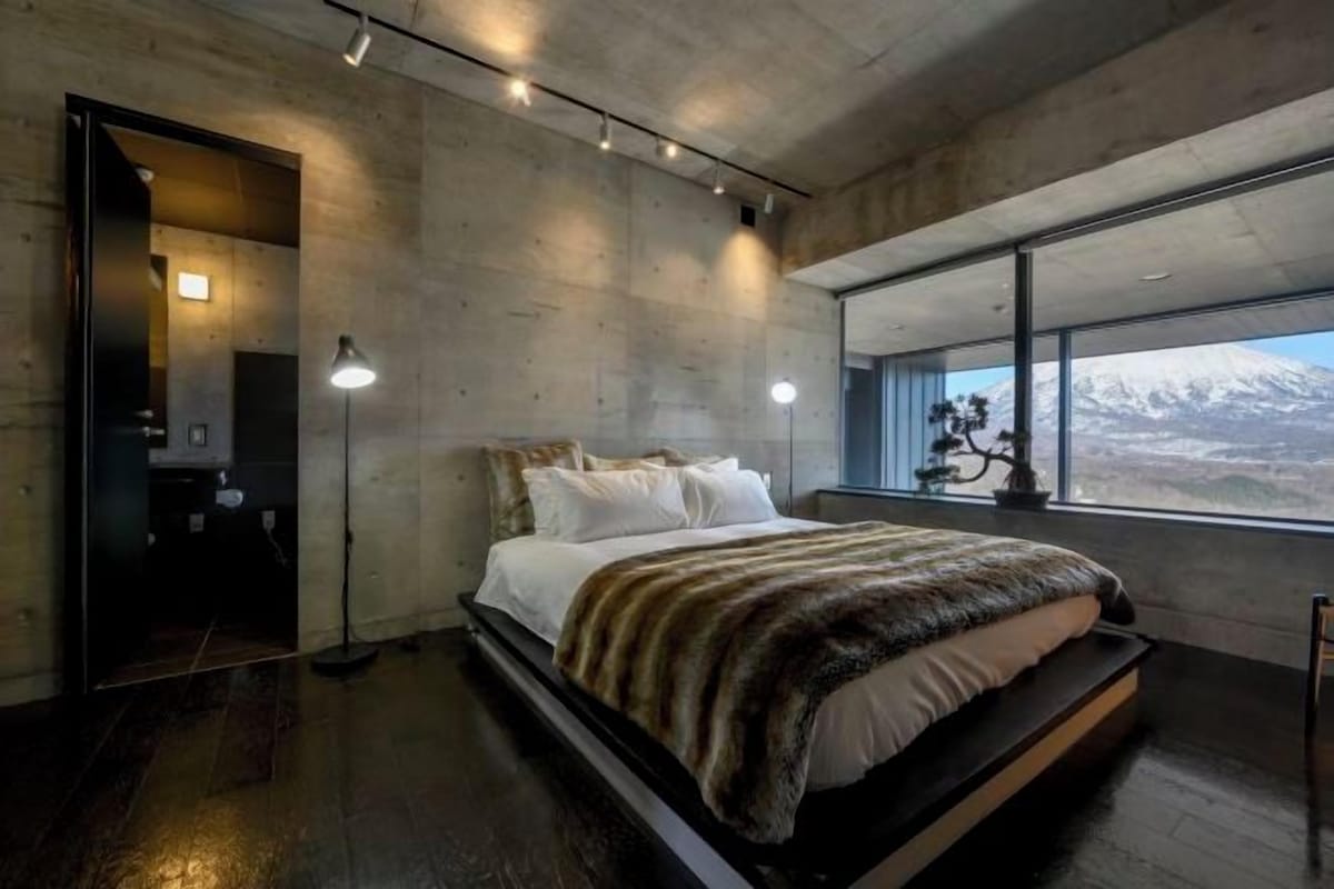 Quarto moderno com cama grande, vista para uma montanha nevada e teto de concreto.