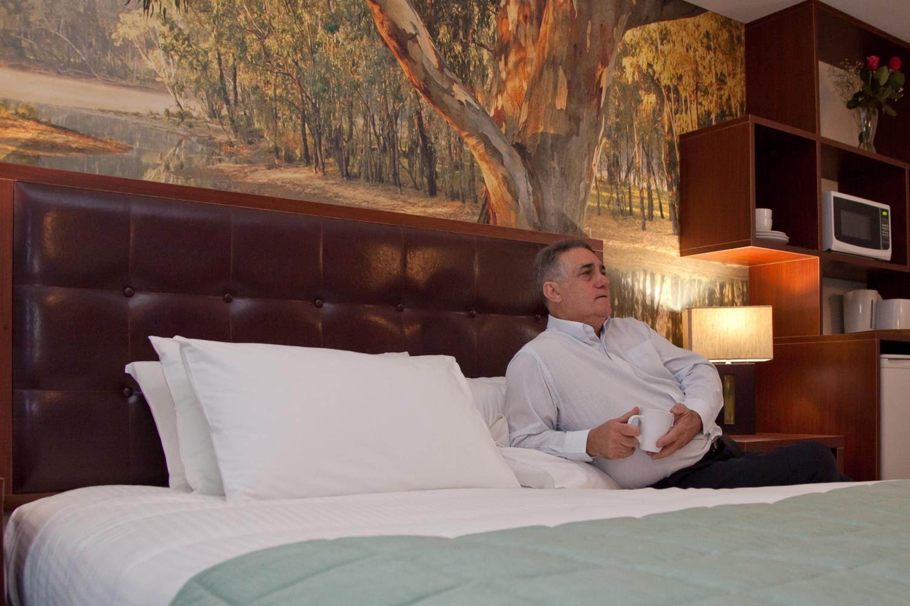 Een hotelkamer met een groot bed, een hoofdbord, een nachtkastje, een lamp, een magnetron en een schilderij aan de muur.