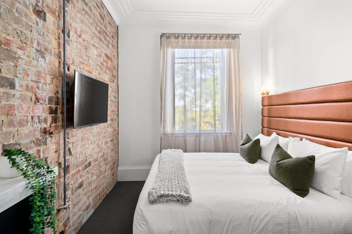 Habitació d'hotel amb paret de maó, un televisor de pantalla plana muntat, un llit gran amb roba de llit blanca i almoades verdes, un reposacapses de fusta i una finestra amb cortines primes.