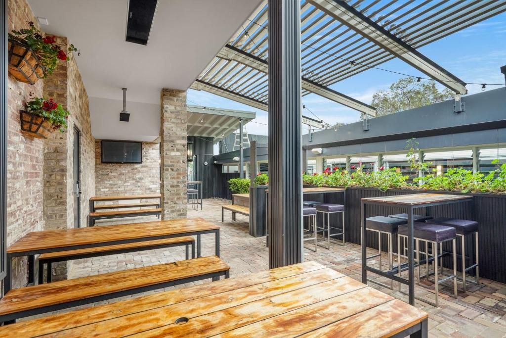 An outdoor seating area with wooden tables and benches, a brick wall, and a glass roof structure.