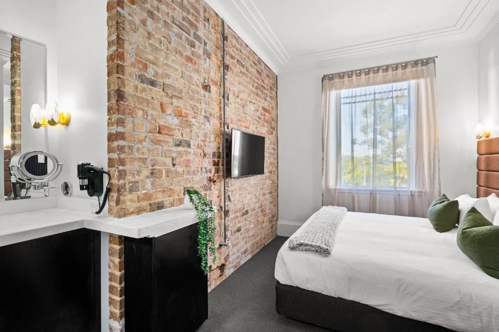 A hotel room with a brick wall, a flat-screen TV, a bed with pillows, a window with curtains, and a bathroom with a mirror and sink.