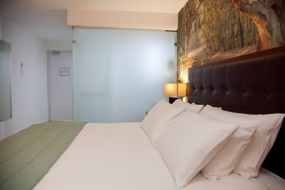 Habitació d'hotel amb un llit gran, un capçal, una làmpada de paret i una porta amb interruptor de llum.