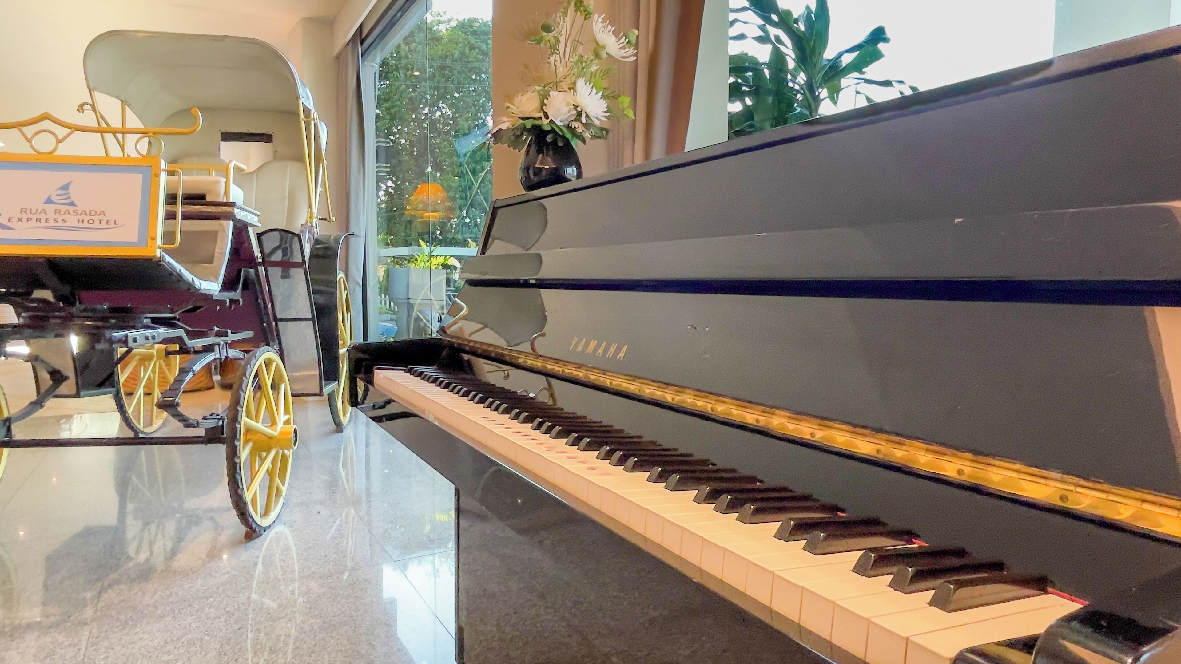 A grand piano in a well-lit room with a glass wall, a vintage carriage, and a sign for Rua Rasada Express Hotel.