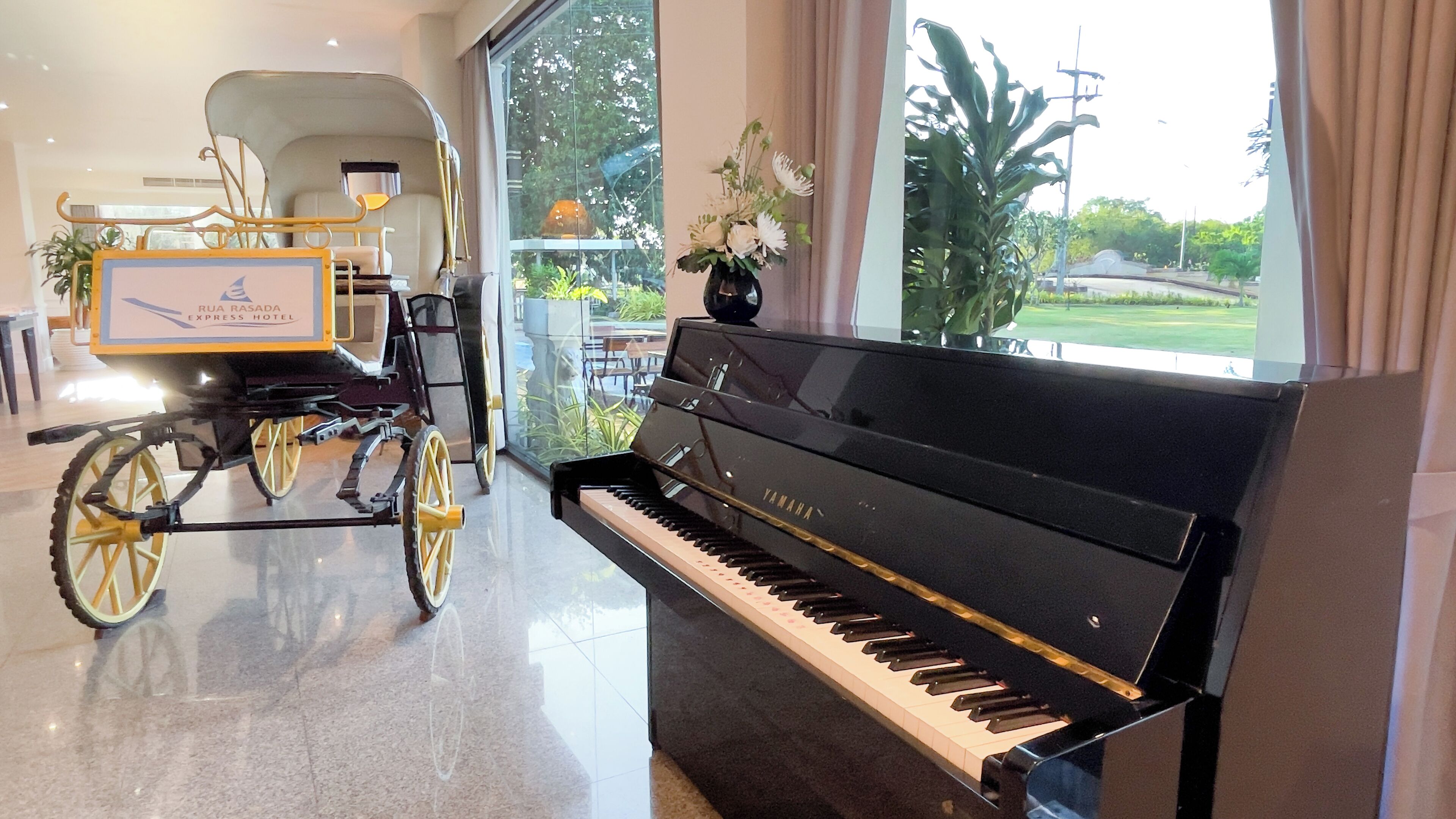 Un piano de cola en una habitación luminosa, con un carruaje y un amplio ventanal que ofrece vistas al follaje exterior.
