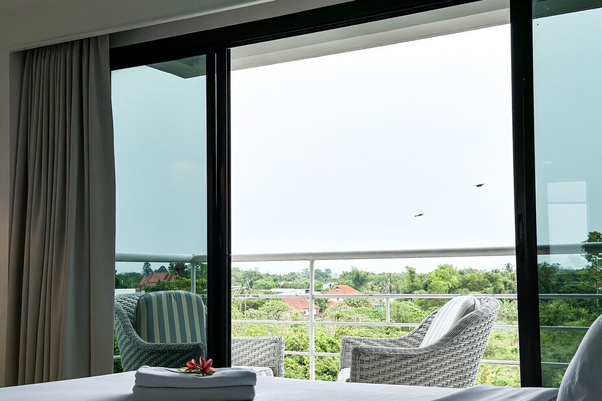 A hotel room with a large window, a bed, a chair, and a balcony with a view of greenery and buildings.