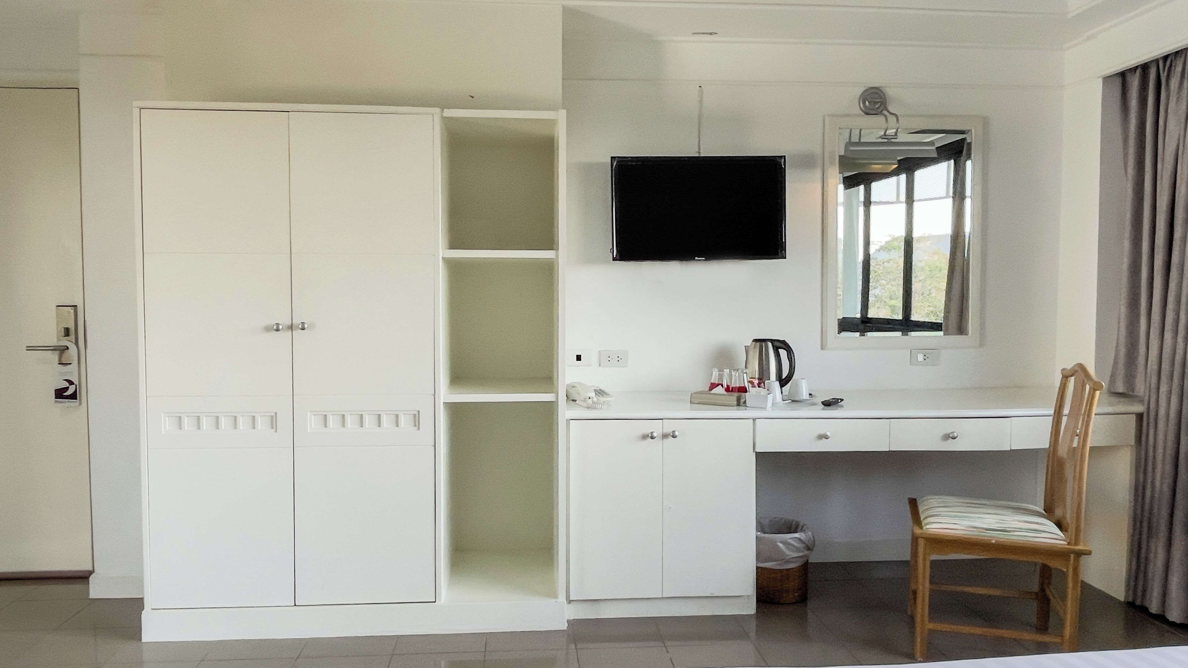 Una habitación con un armario blanco, un escritorio con televisor de pantalla plana, una silla y una ventana con cortinas.