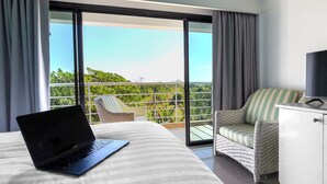 A hotel room with a bed, a laptop, a chair, a TV, and a balcony with a view of greenery. - Rua Rasada Express (Trang)
