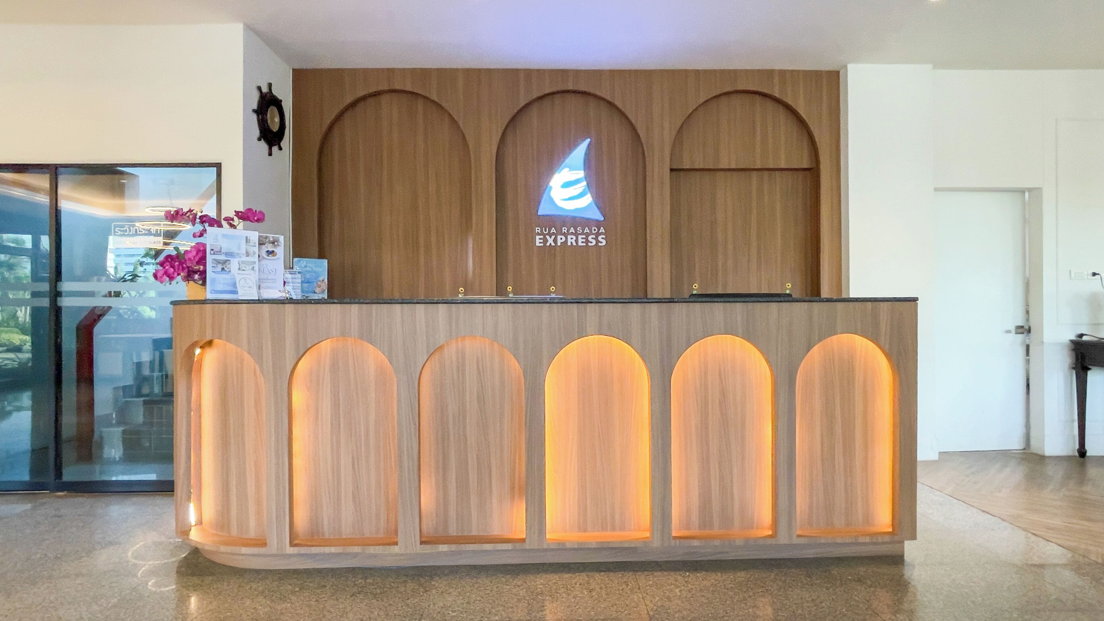 A reception desk with a wooden finish and illuminated panels, a logo above the desk, and a small display of brochures and a vase with flowers.