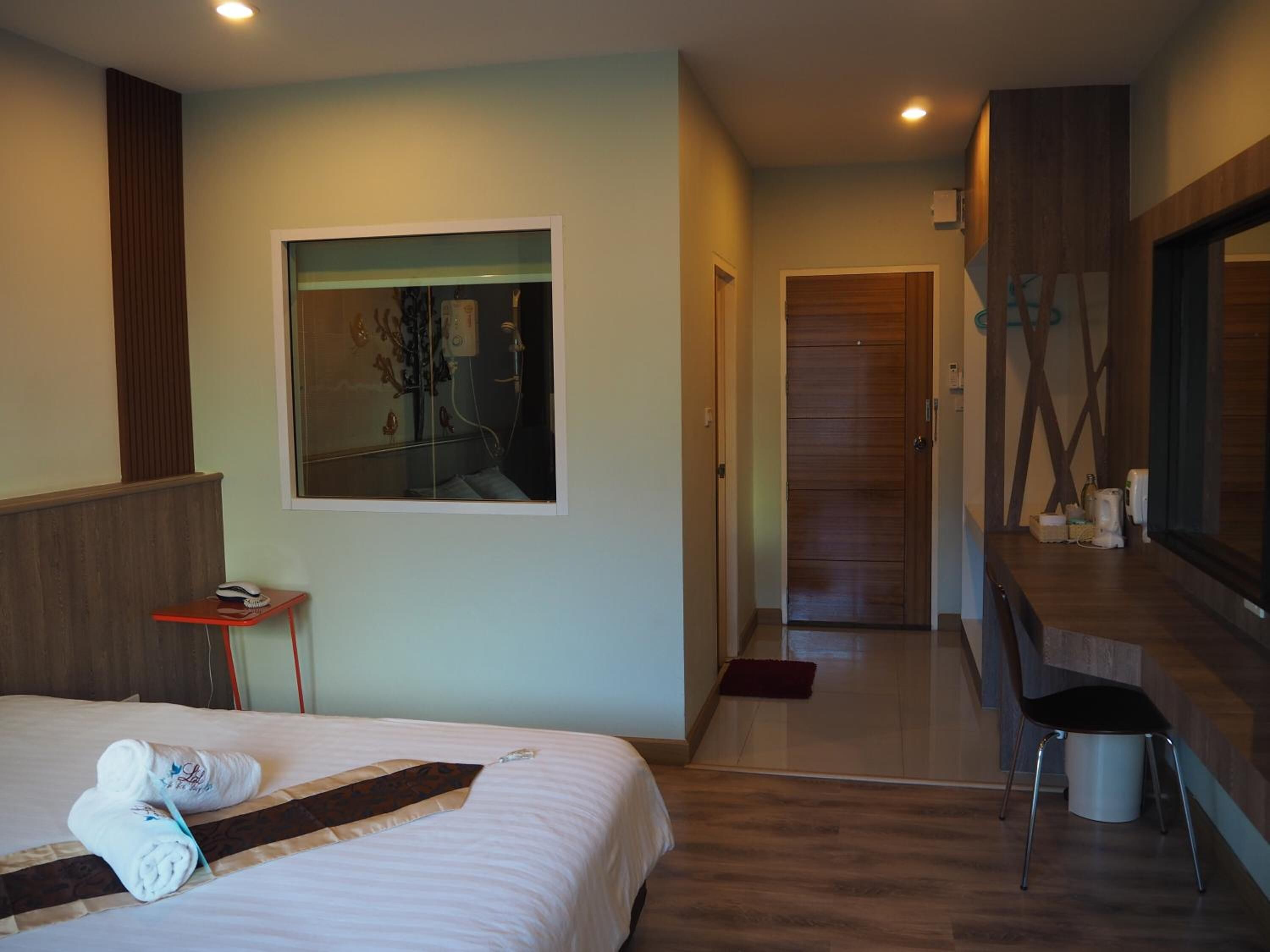 A hotel room with a bed, a small table with a towel, a painting on the wall, and a hallway leading to a bathroom.