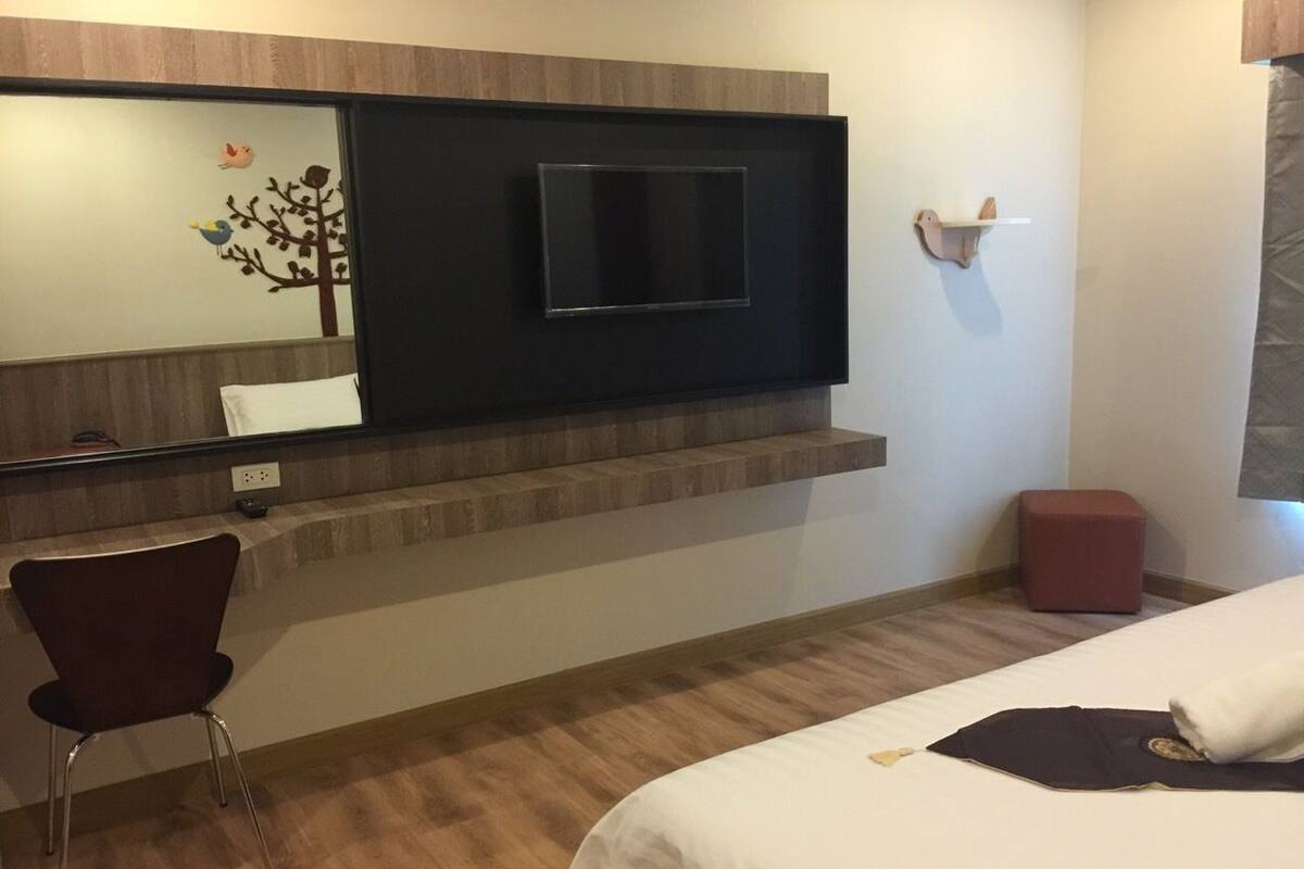 Hotelkamer met een bed, een aan de muur bevestigde televisie, een klein bureau met stoel en een rode poef.