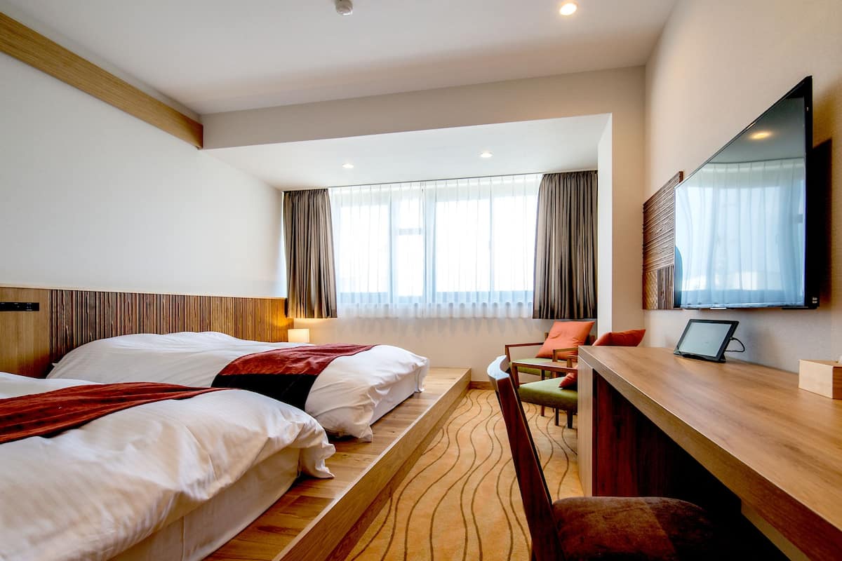 A hotel room with two beds, a desk, a chair, a TV, and a window with curtains.