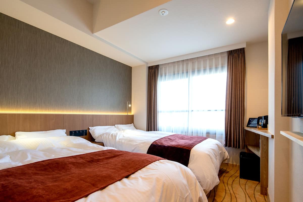 A hotel room with two beds, a large window with curtains, a flat-screen TV, and a small desk with a coffee maker.