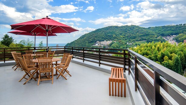 A terrace with wooden furniture and a red umbrella, overlooking a green hillside and a distant town. - Lectore Atami Koarashi (TKP Hotels & Resorts) (Atami)