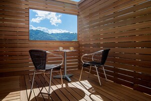 Um deck de madeira com duas cadeiras e uma mesa, uma janela com vista para uma montanha e um céu limpo.