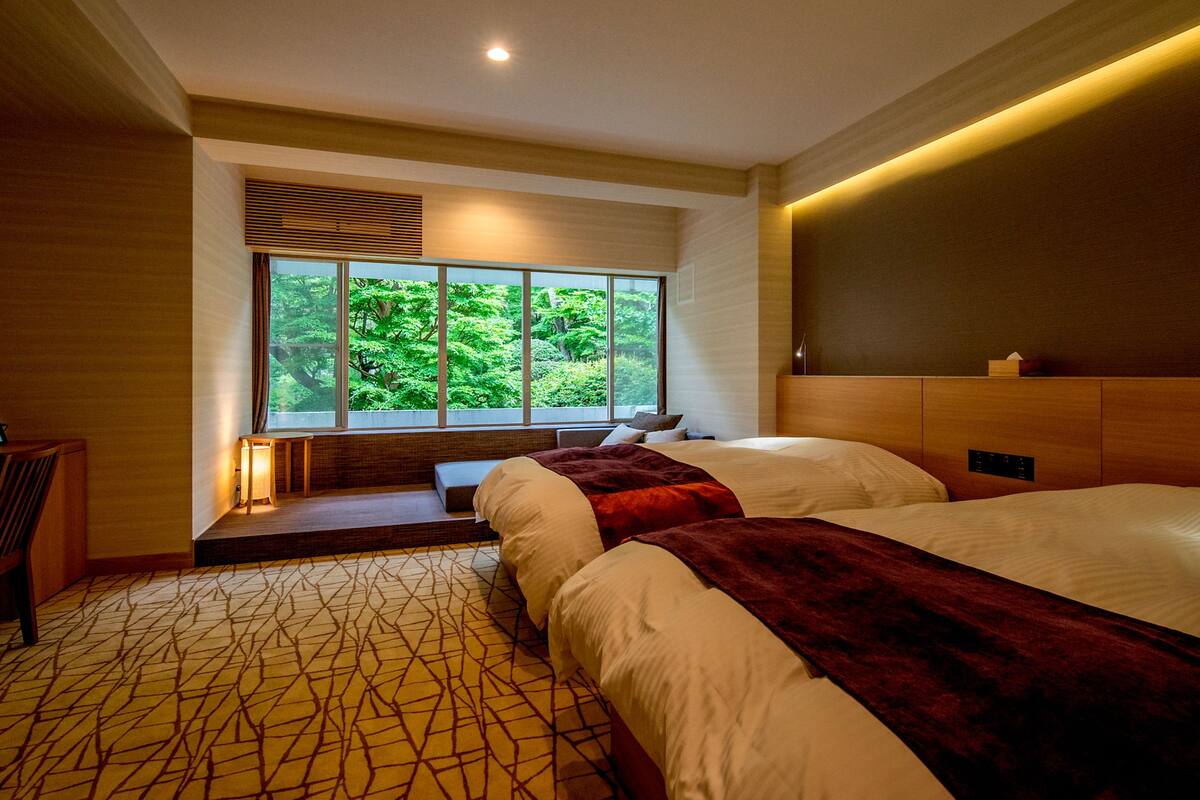 A modern hotel room with two beds, a large window with a view of greenery, a wooden headboard, and a patterned carpet.