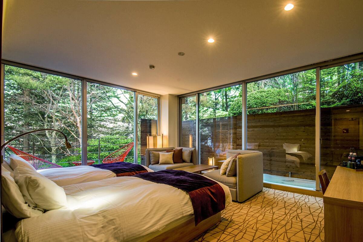 A modern bedroom with a large bed, a seating area with a chair and a table, and large windows offering a view of the outdoors.