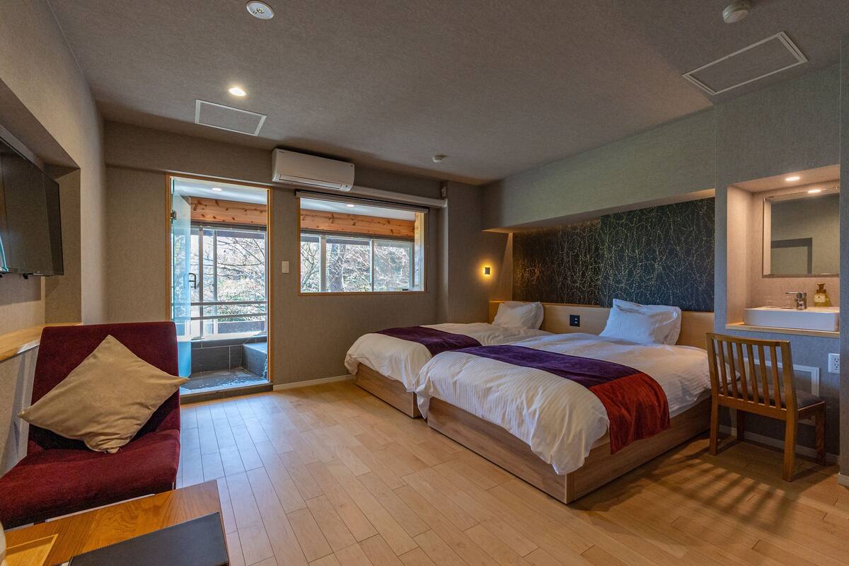 A modern bedroom with a large bed, a seating area with a red chair, a wooden table, and a window providing natural light.
