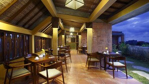 An outdoor dining area with wooden tables and chairs, a brick wall, and a view of the cityscape at dusk. - The Visala Boutique Hotel (Keroboken Kelod)