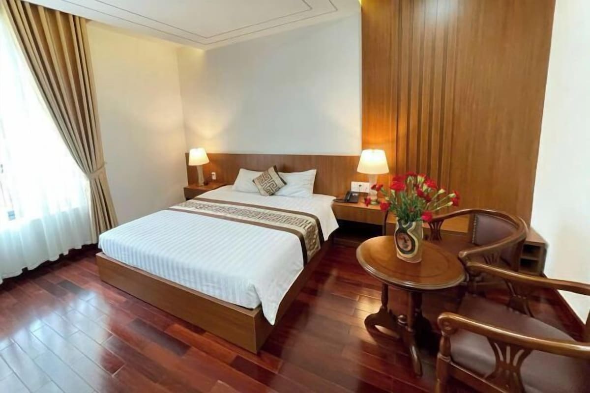 A hotel room with a large bed, two bedside tables with lamps, a wooden chair, and a small round table with a vase of flowers.