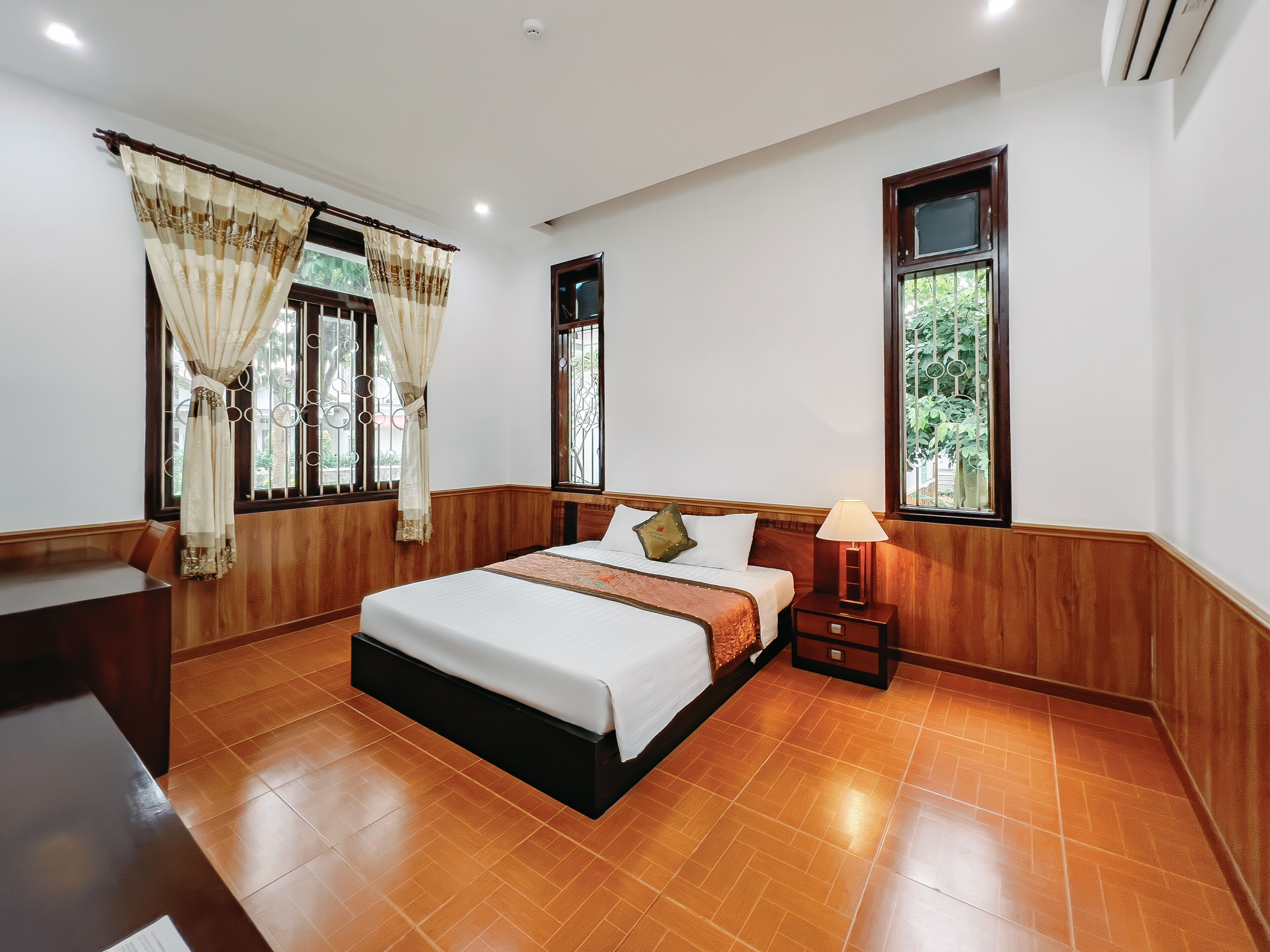 A hotel room with a bed, a nightstand, a lamp, and a window with a view of greenery.