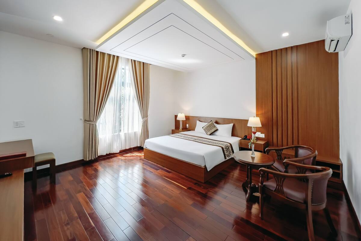 Habitación de hotel con piso de madera, una cama con cabecera blanca, dos mesitas de noche con lámparas, un panel divisor de madera y una ventana con cortinas.