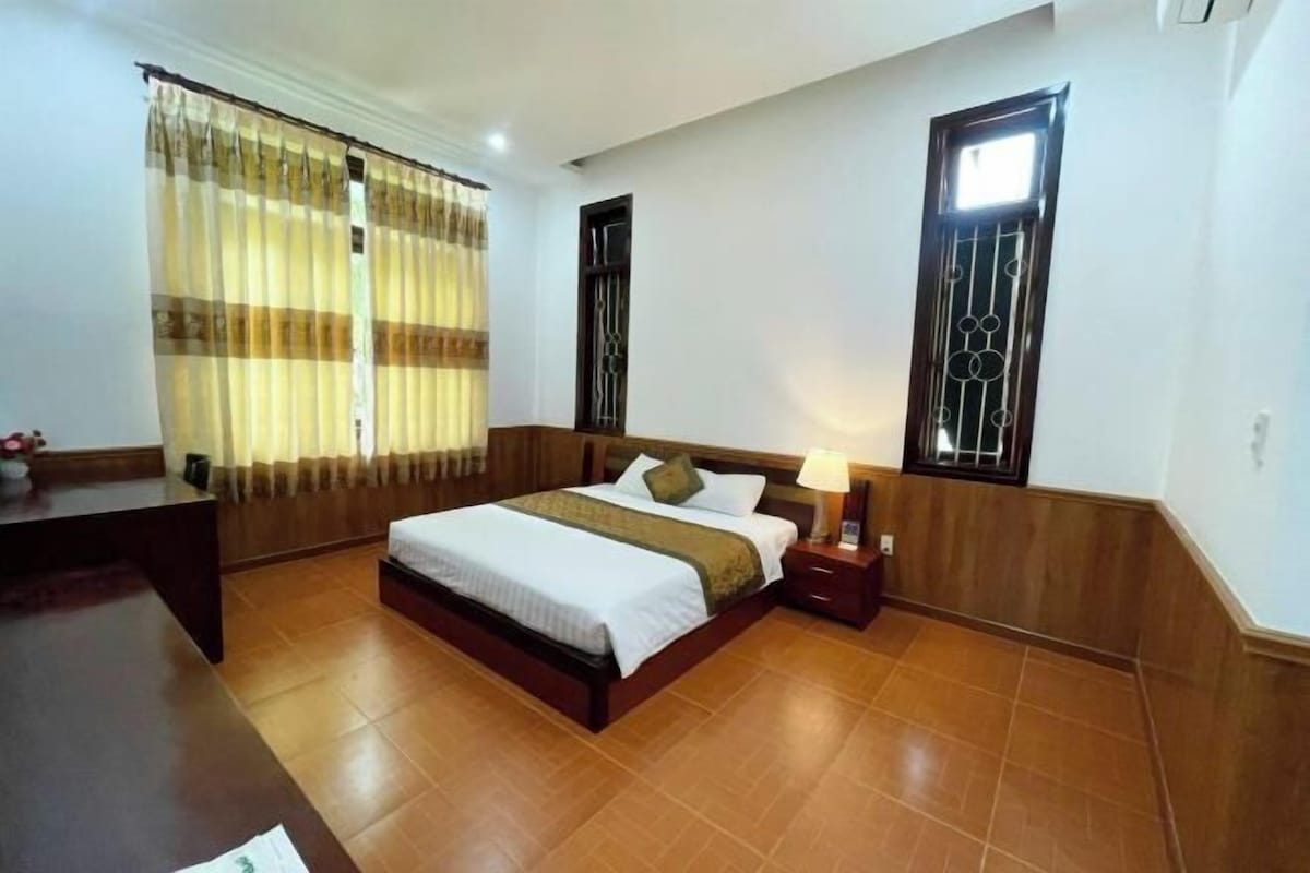 A hotel room with a large bed, wooden headboard, a nightstand with a lamp, a desk, and a window with decorative bars.