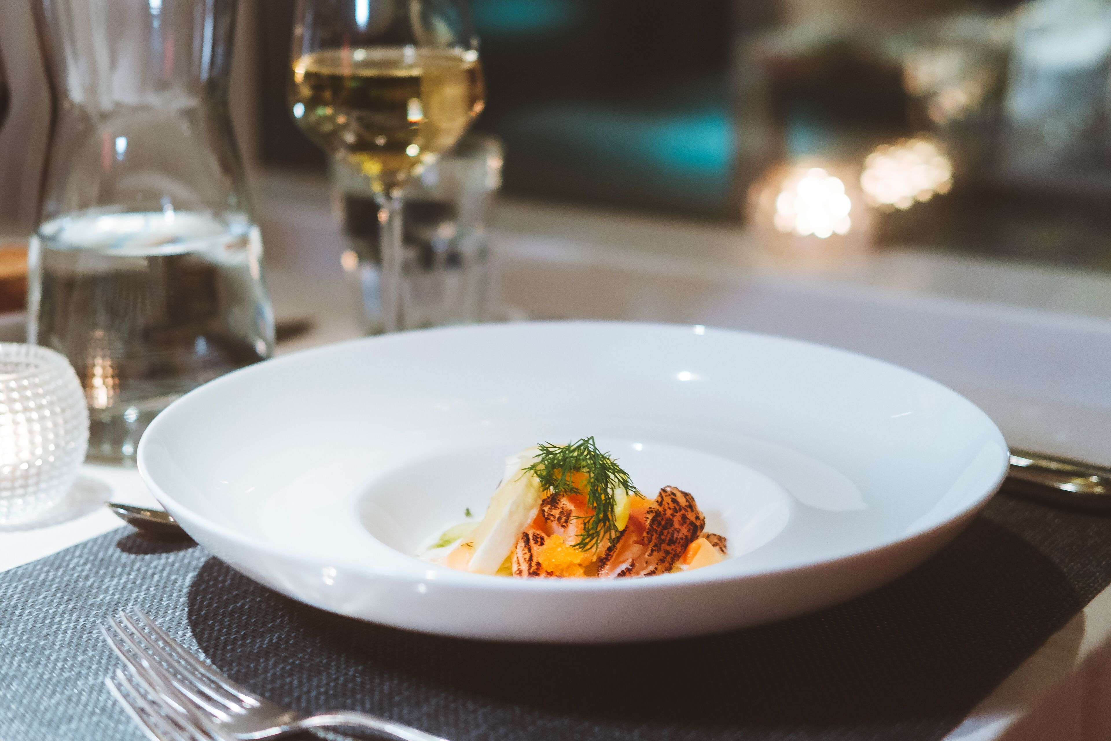A white plate with a dish of food, a fork, and a glass of white wine on a table.
