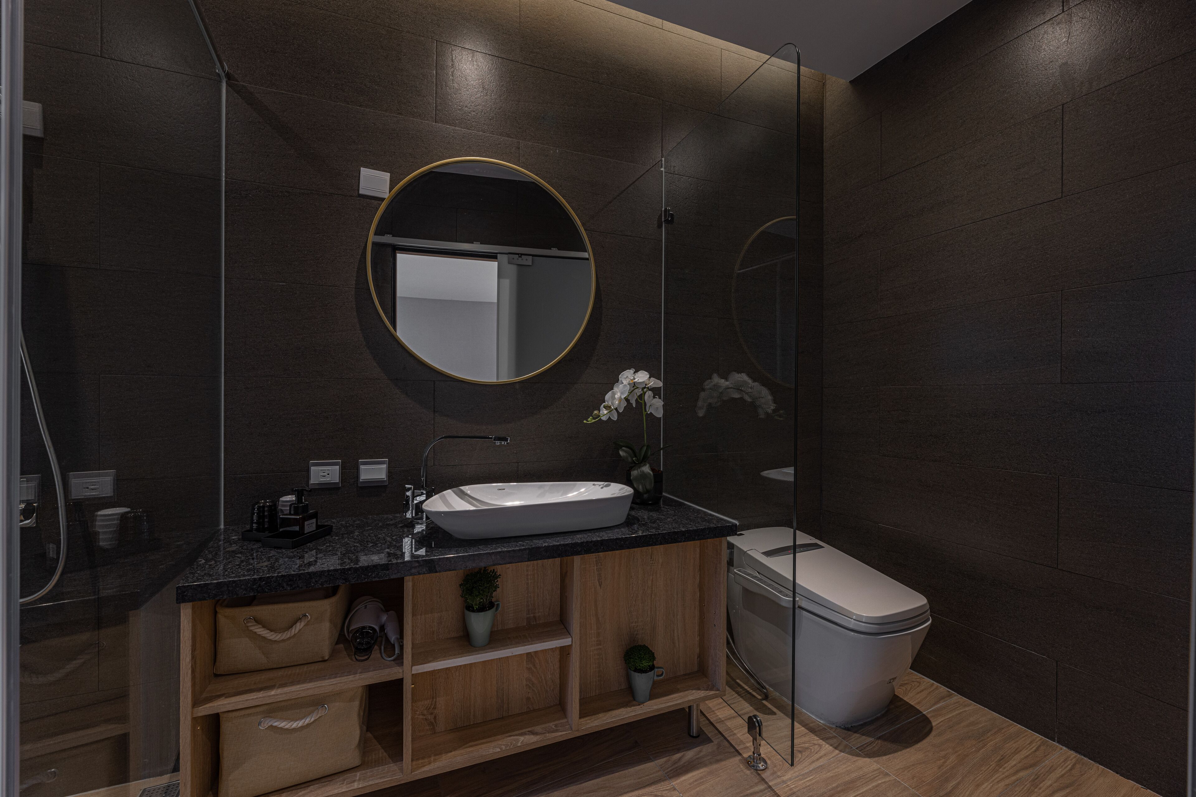 A modern bathroom with a dark wood finish, a white sink, a round mirror, and a toilet.