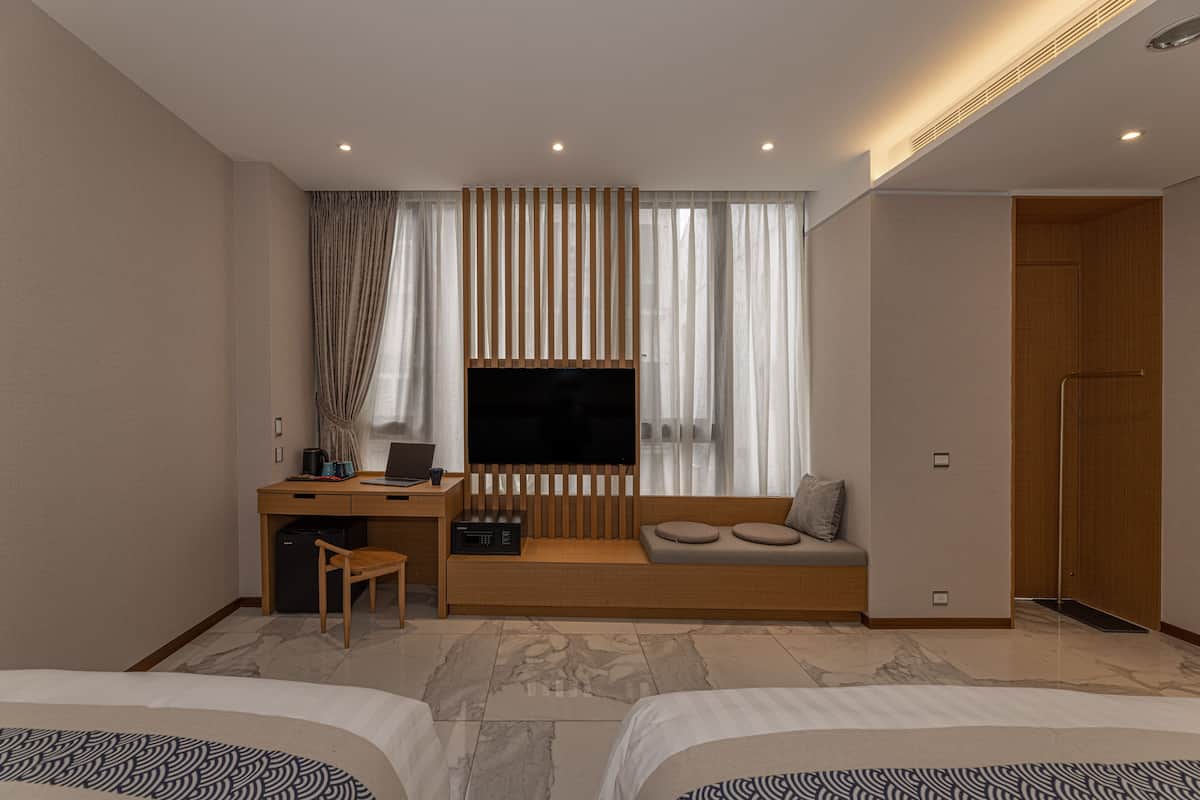 A modern hotel room with a bed, a desk with a computer, a TV, and a seating area with cushions.