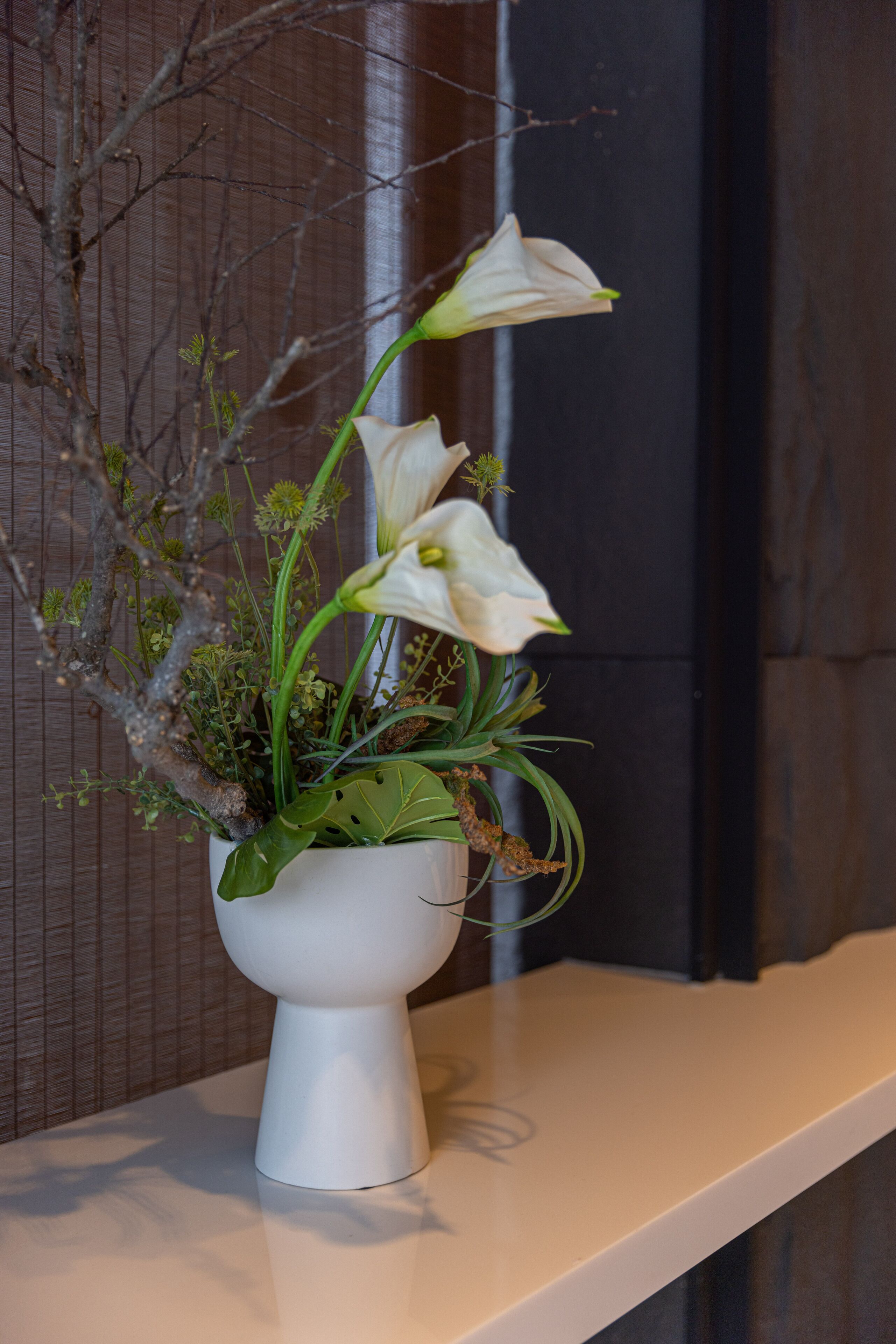 A white ceramic vase with a floral arrangement on a white shelf against a dark wall.