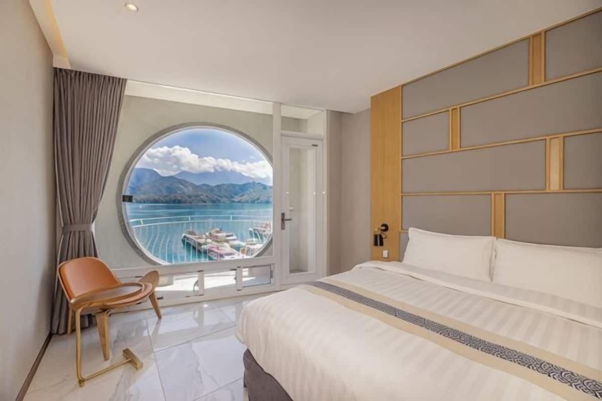 A hotel room with a large bed, a circular window with a view of the sea, a chair, and a door leading to a balcony.
