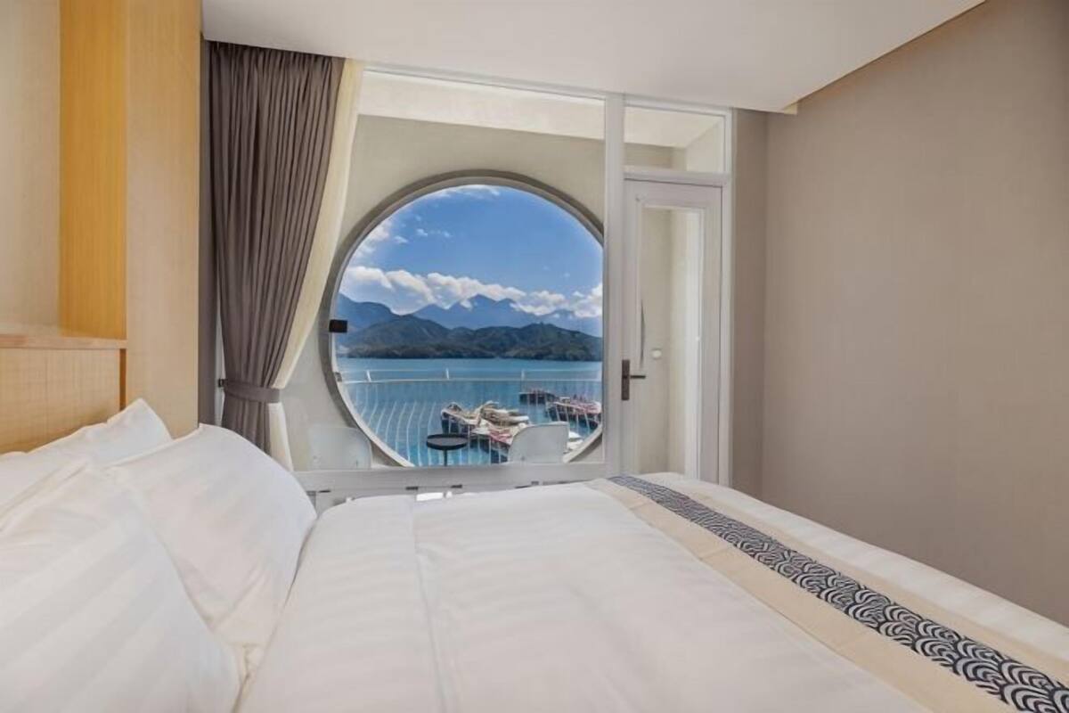 A hotel room with a large bed, a circular window with a view of a pool and mountains, and a balcony with chairs.