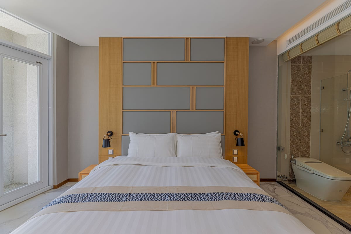 A modern hotel room with a large bed, a wooden headboard, a bedside table with a lamp, and a bathroom with a shower and toilet.