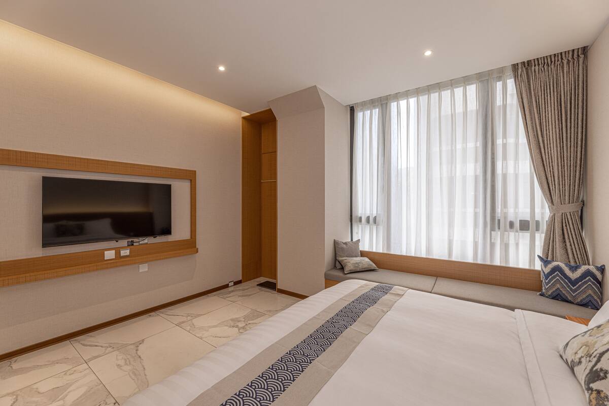 A modern hotel room with a large bed, a flat-screen TV mounted on the wall, a window with sheer curtains, and a built-in bench with cushions.