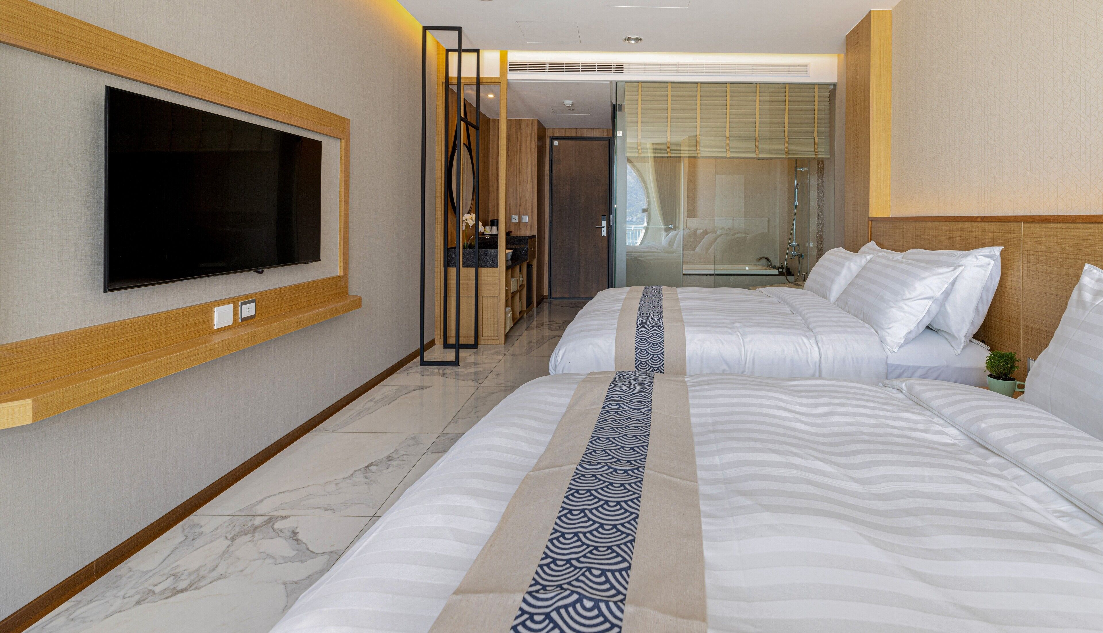 A hotel room with two beds, a TV mounted on the wall, a bathroom with a glass door, and a small plant on the bedside table.
