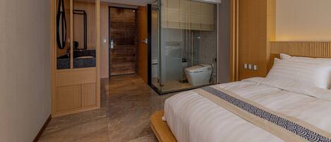 Una habitación de hotel moderna con una cama grande, un vestidor, una ducha con mampara de vidrio y un cabecero de madera.