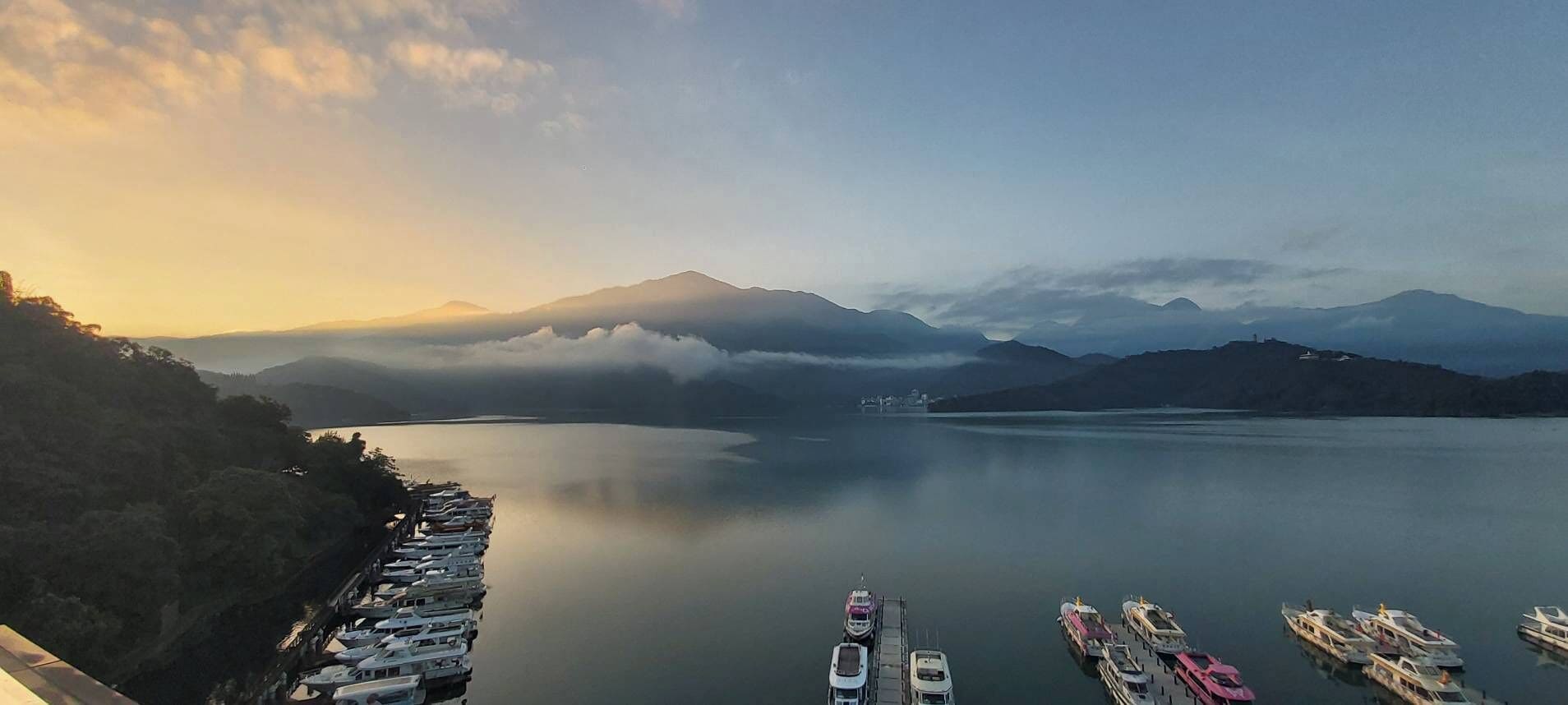 Un port de plaisance où sont amarrés des bateaux, entouré de montagnes et bordé par une étendue d’eau.