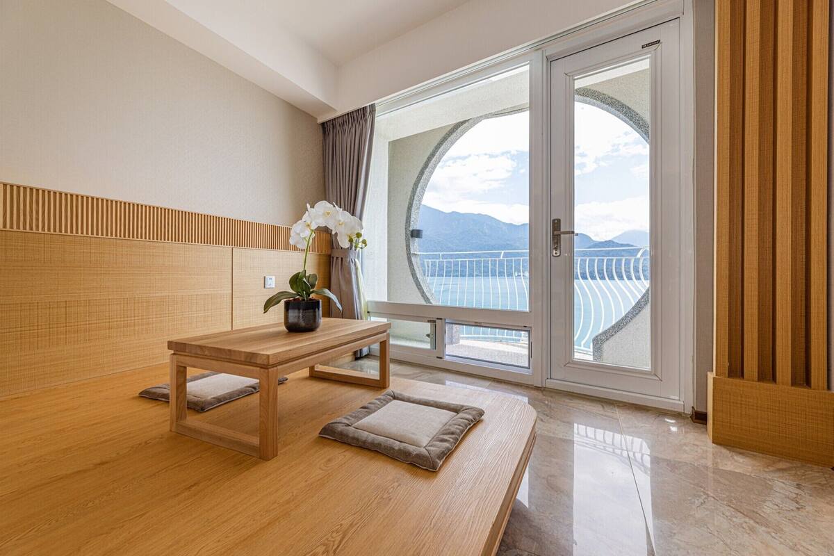 A modern room with a wooden floor, a small table with a cushion, a potted plant, and a view of a balcony with a railing and mountains in the background.