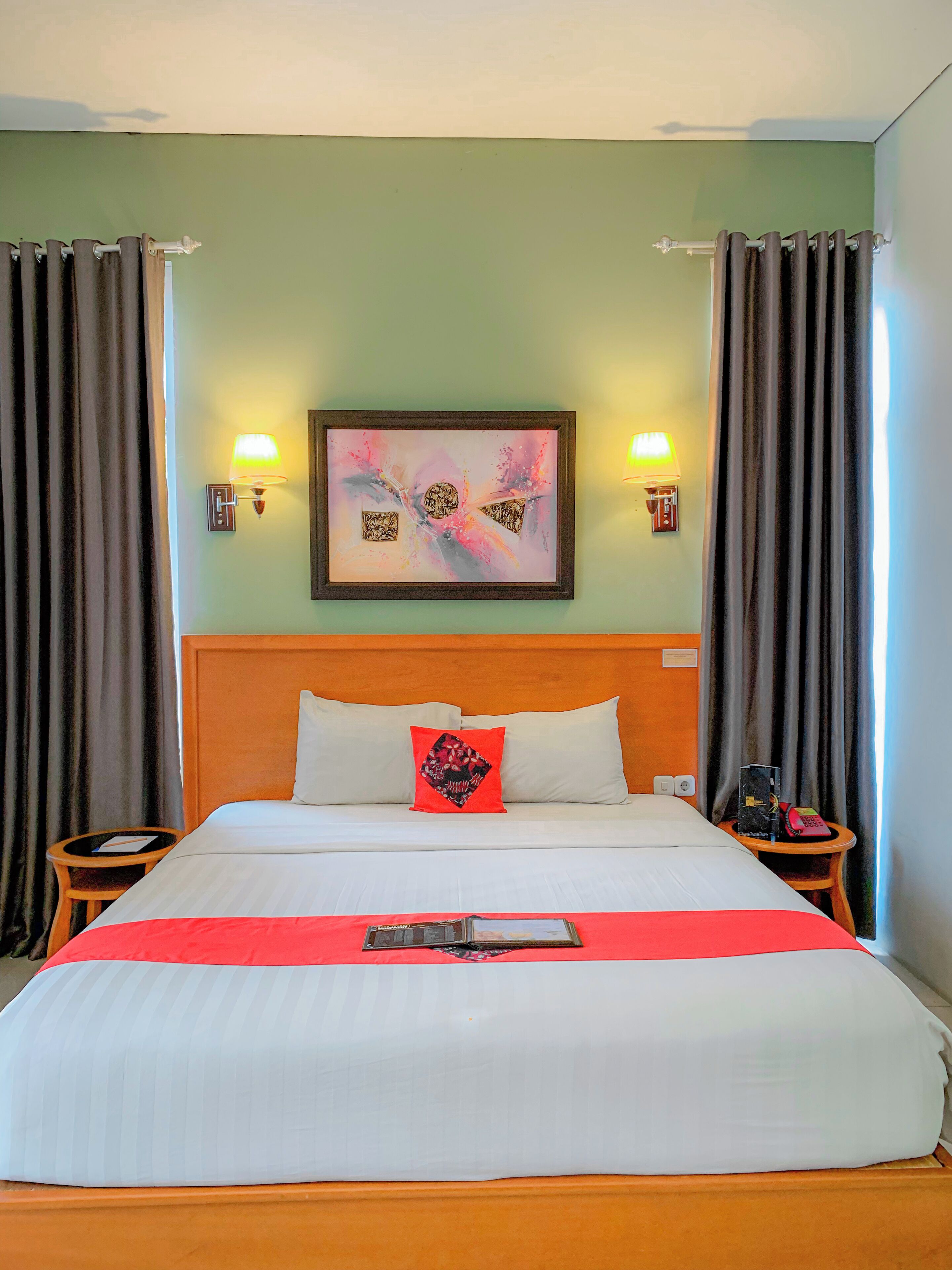 A hotel room with a large bed, two bedside tables, a headboard, a framed abstract painting, and two wall-mounted lamps.