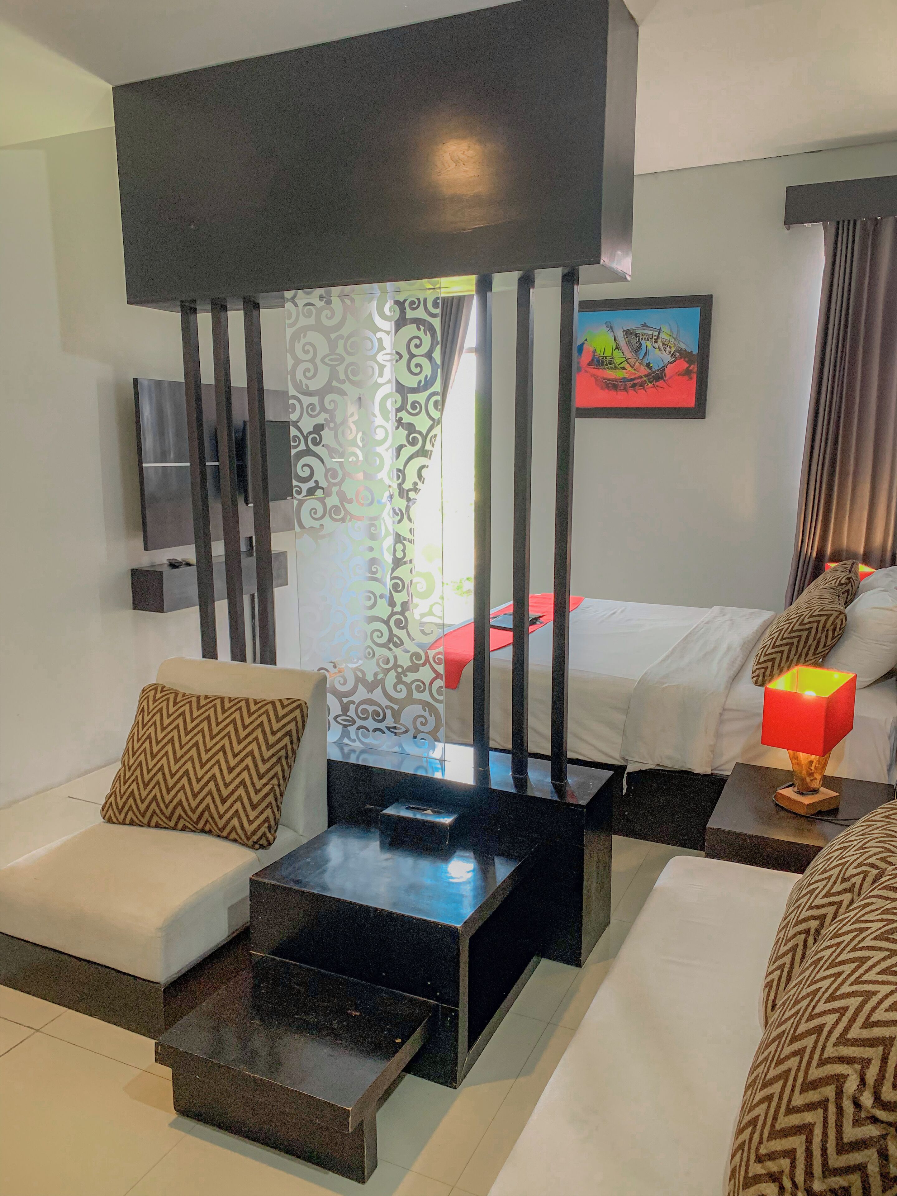 A modern hotel room with a sofa, a coffee table, a bed with a red blanket, a painting on the wall, and a TV mounted on the wall.