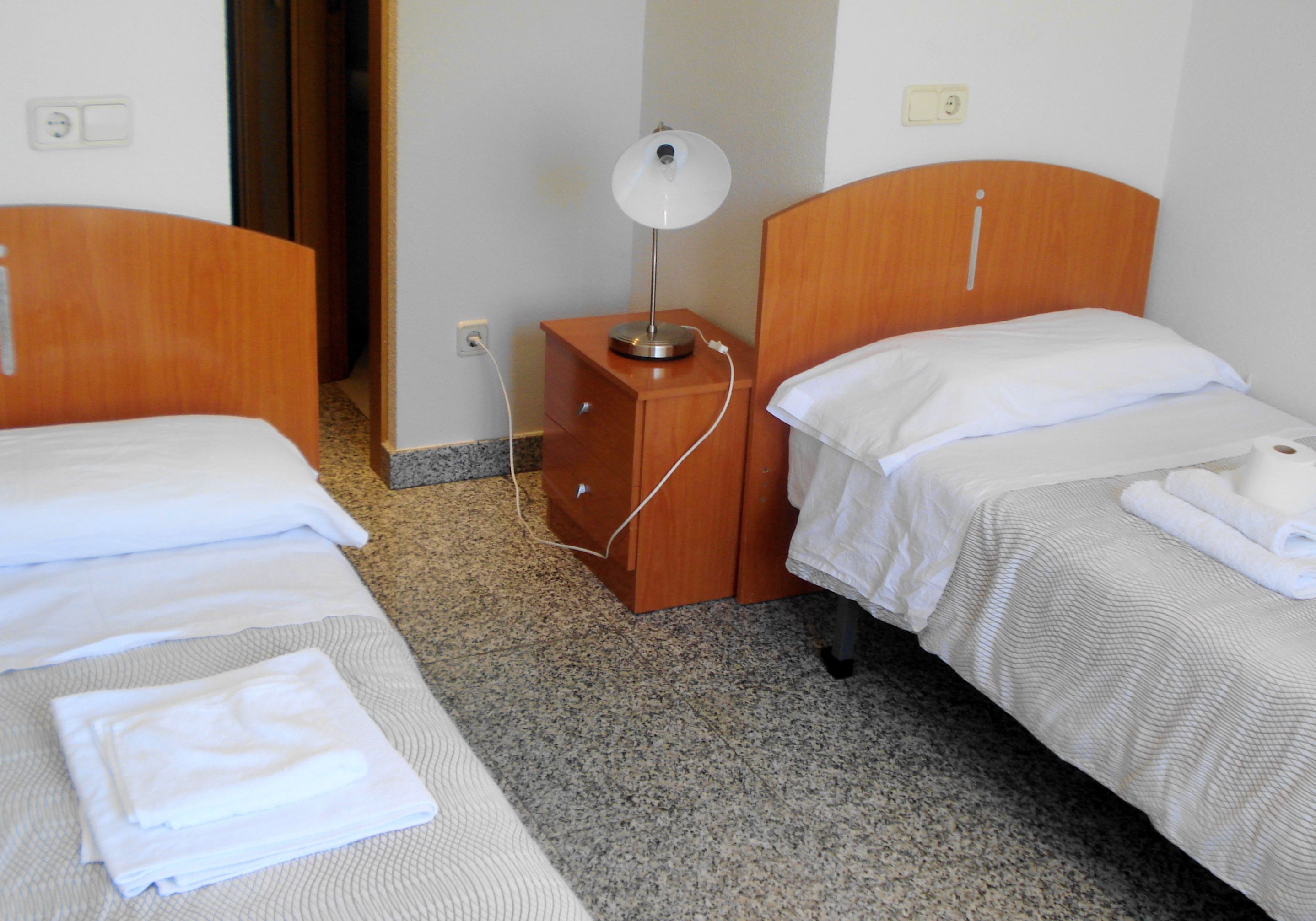 Une chambre d’hôtel avec deux lits simples, une table de chevet en bois avec une lampe et un petit tiroir.